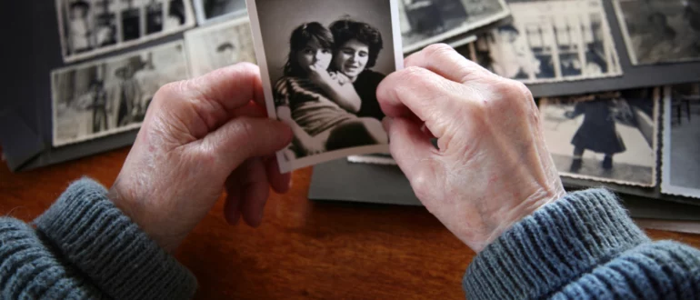 Руки пожилой женщины держат черно-белую фотографию молодой матери и ребенка с фотоальбомами на заднем плане.