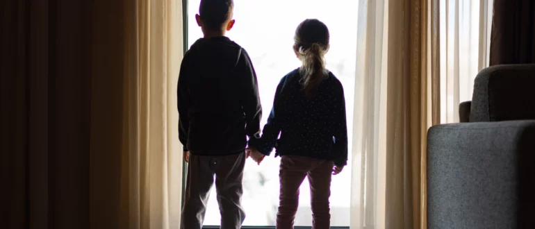 Силуэт молодых братьев и сестер, держащихся за руки и смотрящих в окно.