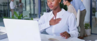 Poklicna ženska sedi za pisalno mizo z zaprtimi očmi in drži eno roko na prsih, medtem ko njeni sodelavci delajo v ozadju.