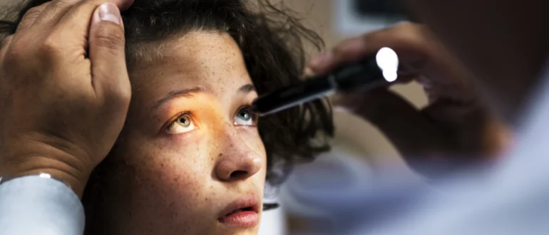 Выборочный фокус на девочке-подростке, смотрящей вверх, когда врач светит лампой, осматривая ее глаза.