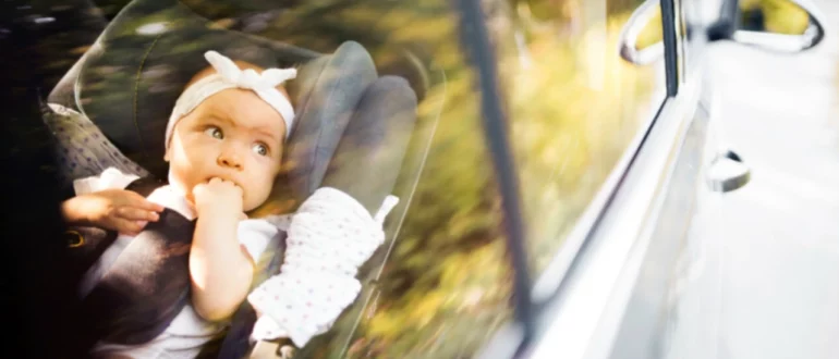 Egy autó ablakából nézve készült fotó, amelyen egy kislány az ujjait szopogatja, miközben egyedül van bekötve az anyósülésbe.