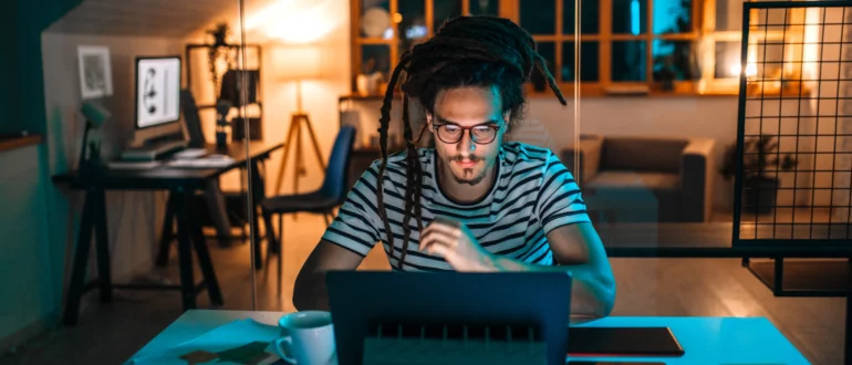 Egy szemüveges férfi egy laptopon dolgozik egy íróasztalnál éjszaka egy irodai környezetben.